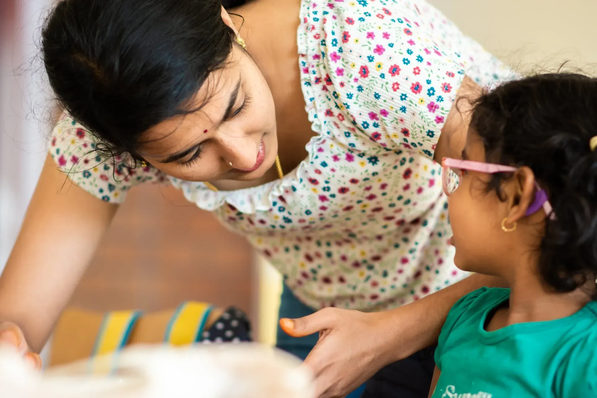 A teacher leaning in to talk warmly with a young girl wearing glasses