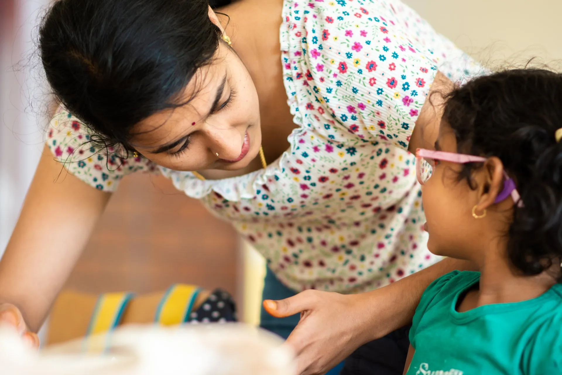 A teacher leaning in to talk warmly with a young girl wearing glasses