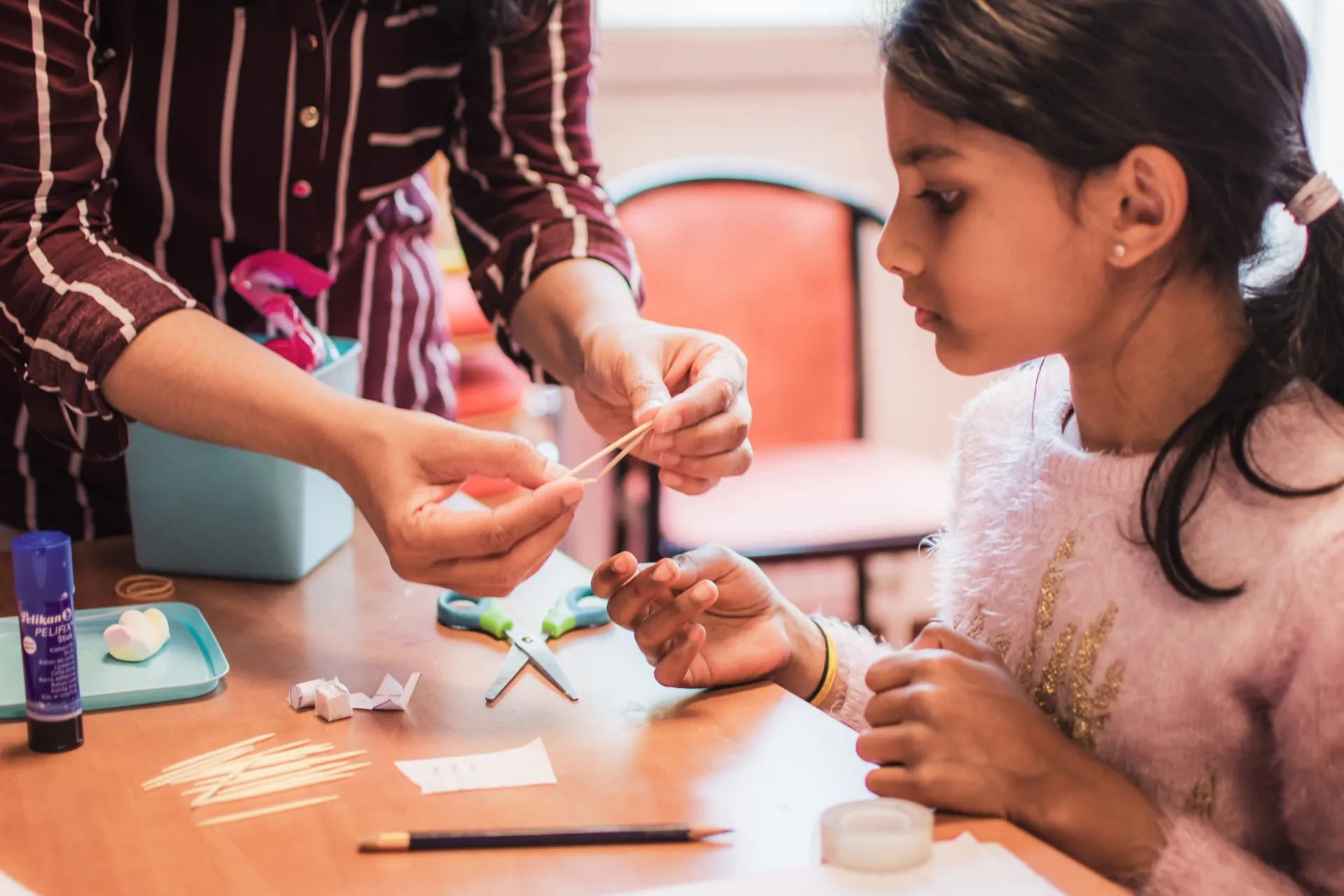 A teacher helping a girl with a hands-on craft activity using sticks and glue
